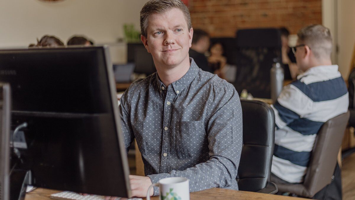 A man (James) sat at a desk on his computer, smiling at the camera.