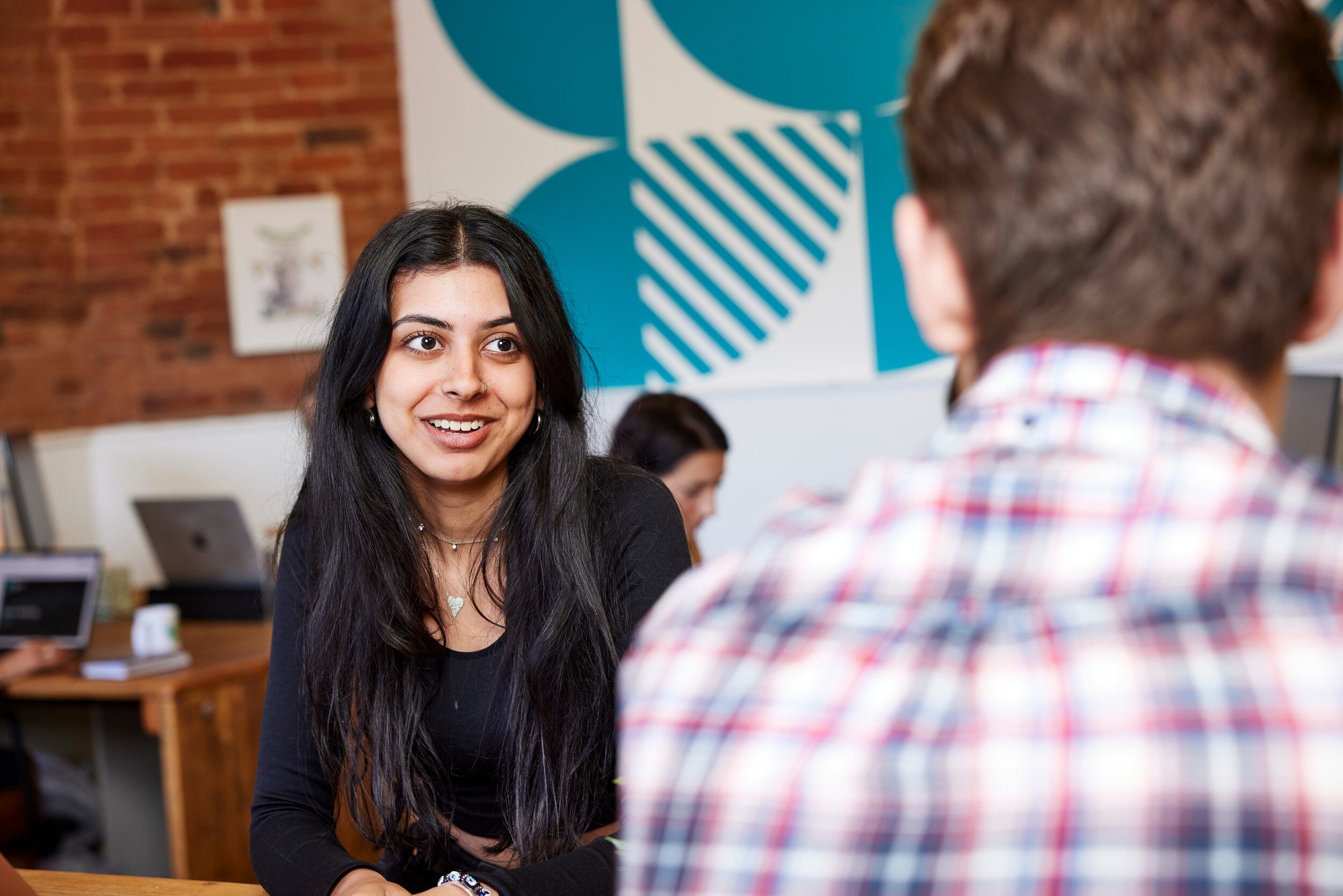 Girl smiling at a boy sitting at a table in an office, with his back to the camera.